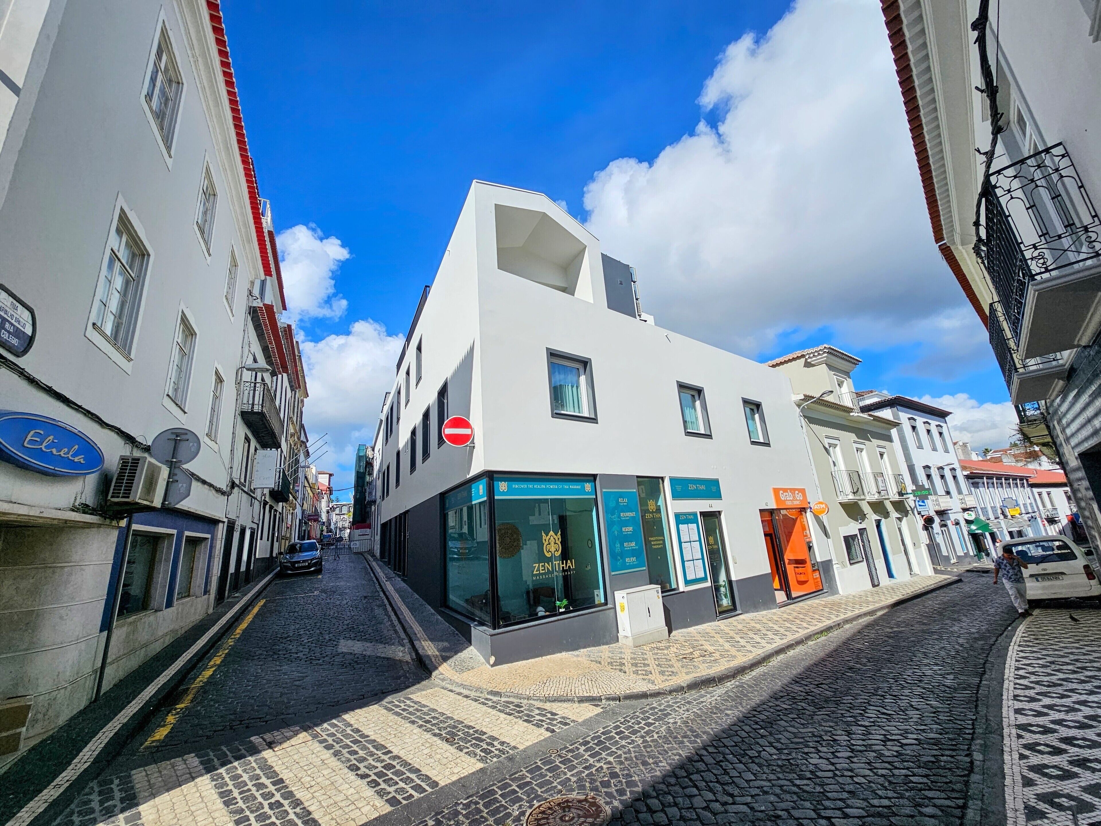 Una vista urbana amb un edifici blanc que presenta un restaurant Zen Thai, un rètol blau per Eliel, i un carrer pavimentat de pedra.