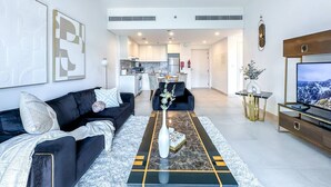 A modern living room with a black sofa, a coffee table, and a TV. There is a kitchen in the background. - UltraLuxury Flat Amazing Pool Near to Burj Al Arab (Jumeirah Medinat Living)