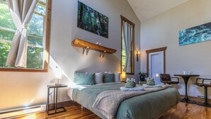 A bedroom with a large bed, wooden flooring, a window with curtains, a bedside table, a nightstand, and a wall-mounted shelf.
