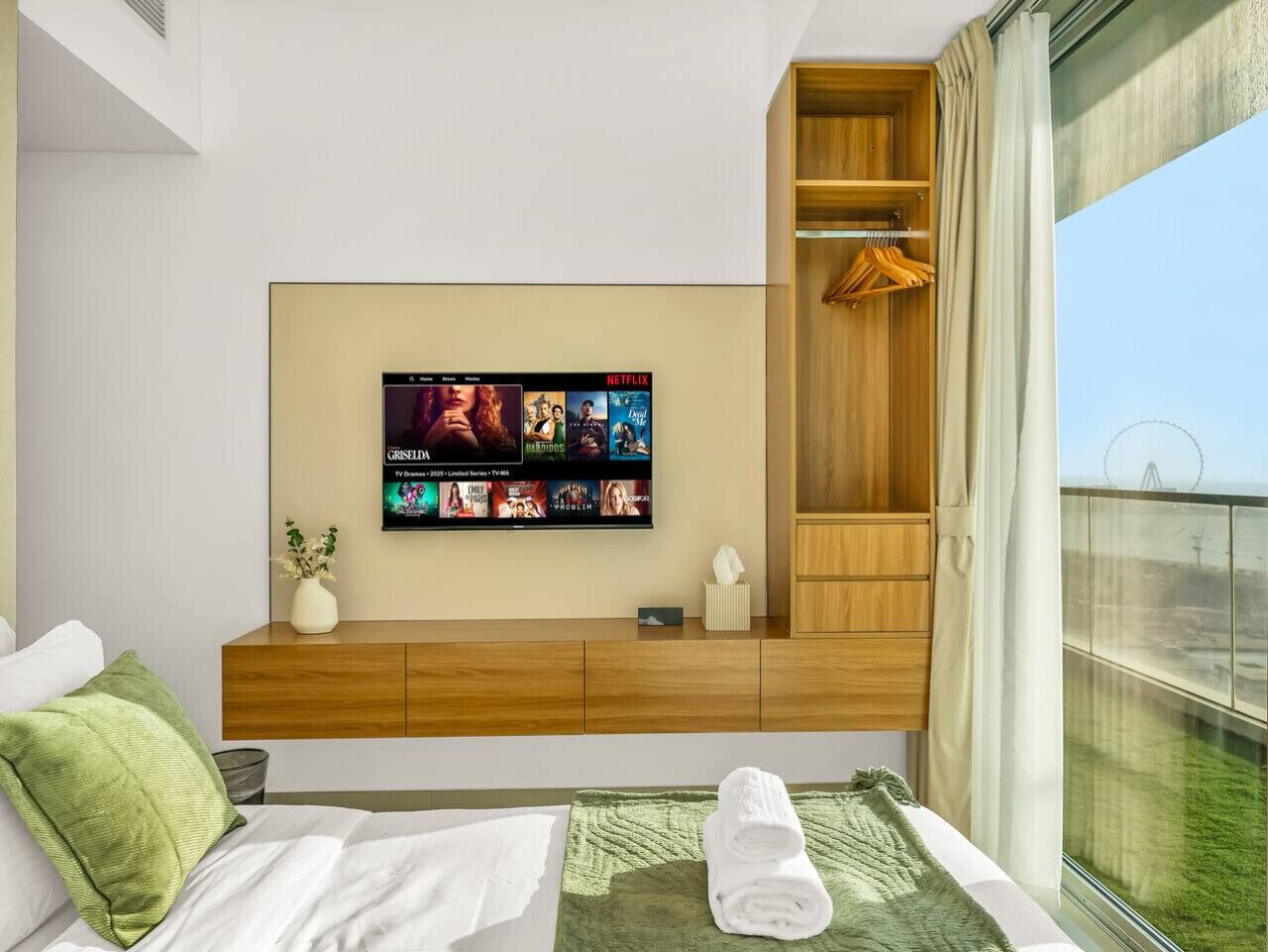 A modern hotel room with a large flat-screen TV mounted on the wall, a wooden console table, a bed with white linens, a green throw blanket, and a view of a Ferris wheel through a large window.