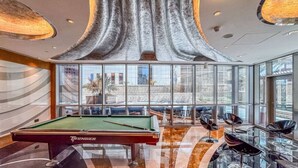Une table de billard recouverte d’un drap vert, avec un queue de billard à proximité, entourée de tabourets de bar modernes et d’un sol incurvé réfléchissant.