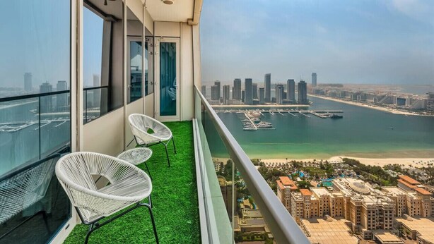 Un balcon avec deux fauteuils en osier blancs, une porte vitrée et une vue sur les toits d’une ville et la côte.