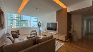 A modern living room with a large sofa, a flat-screen TV, wooden paneling, and a large window with a view of buildings. - Opulent Flat with Beach, Pool & Canal View in JBR (Dubai Marina)