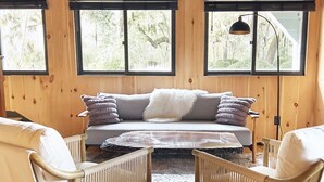 A cozy cabin interior with a wooden floor, a sofa with cushions, two wicker chairs, and large windows offering a view of the forest. - Wicklow Hall (Georgetown)