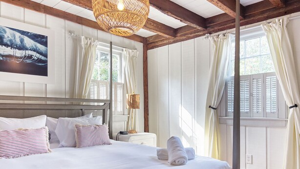 A neatly made bed with white linens and striped pillows, a wicker lamp, and a view of the outdoors through a window with white curtains. - Wicklow Hall (Georgetown)