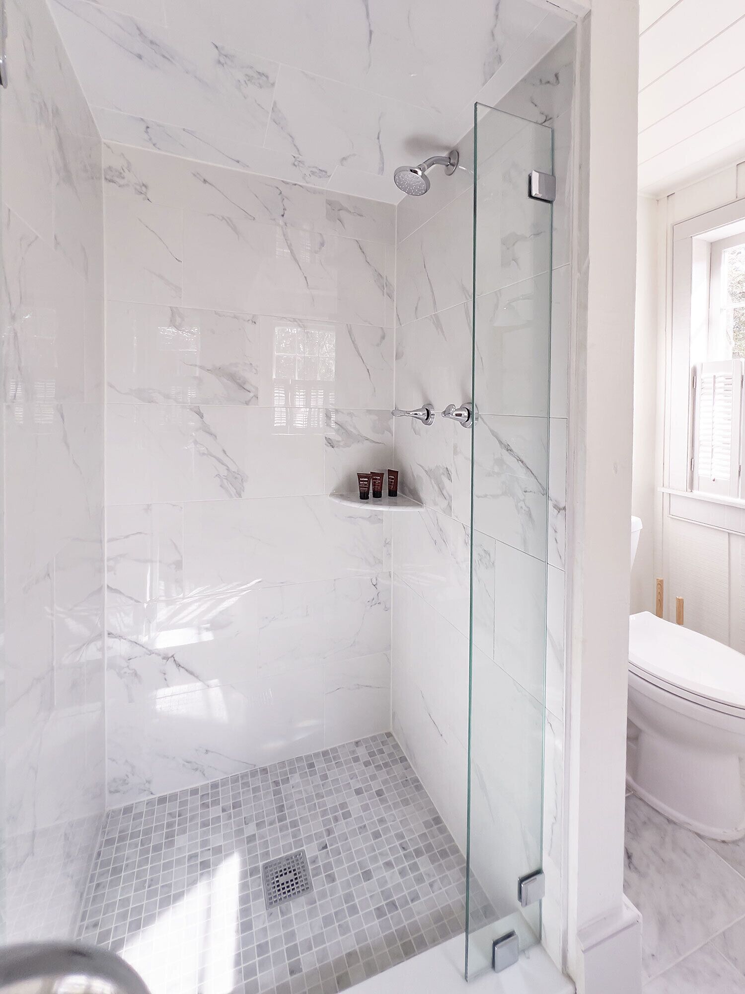 Een moderne badkamer met een douche met glazen wand, marmeren wanden en een wit toilet.