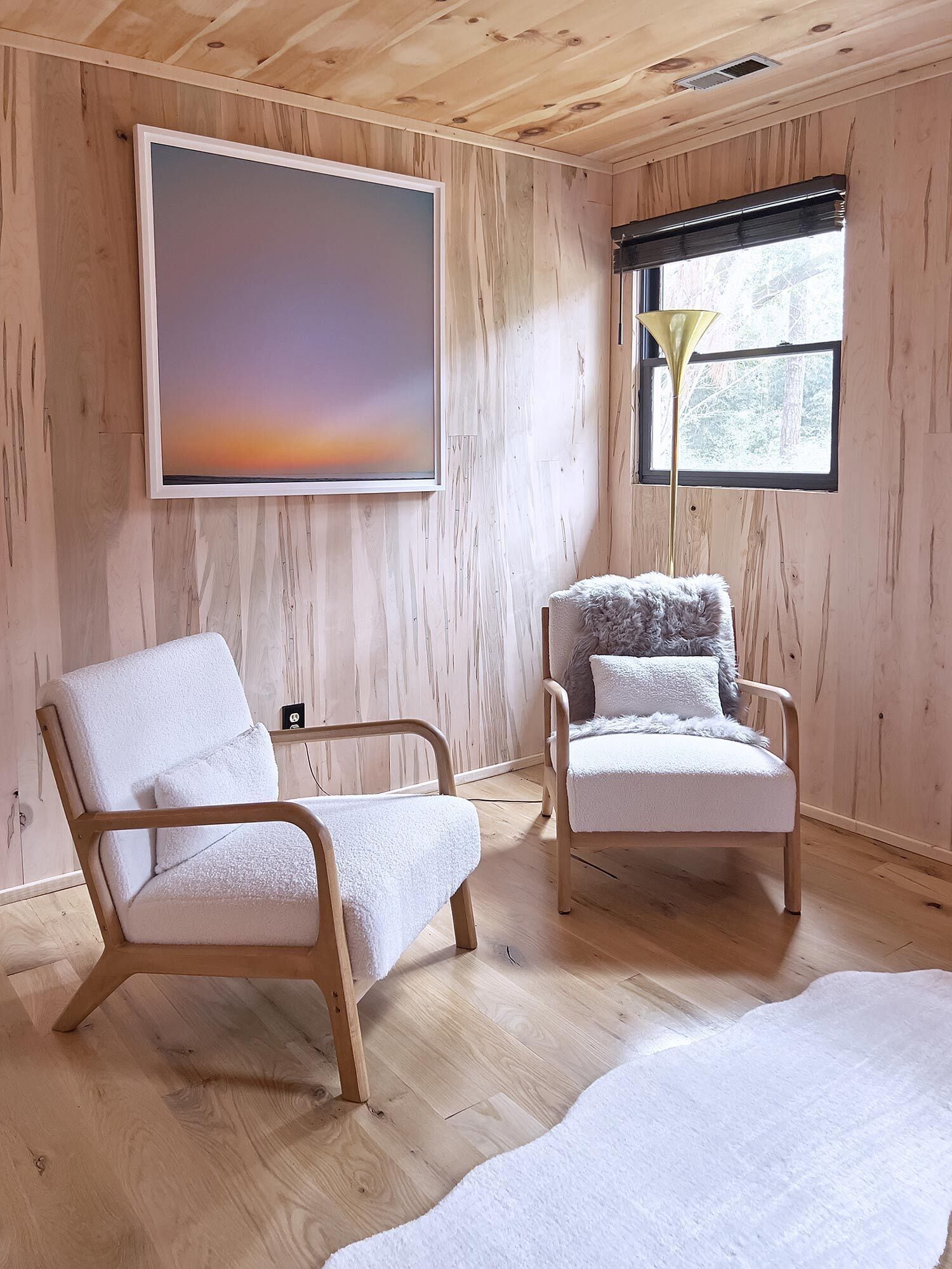 A room with wooden walls and floors, two white chairs, a large abstract painting, and a window with a view of trees.
