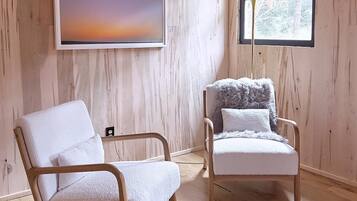 Ein Zimmer mit Holzwänden und -böden, zwei weiße Stühle, ein großes abstraktes Gemälde und ein Fenster mit Blick auf Bäume.