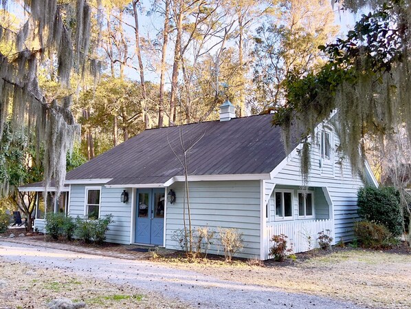 A small, single-story house with a dark roof and white siding, surrounded by trees and a gravel driveway.