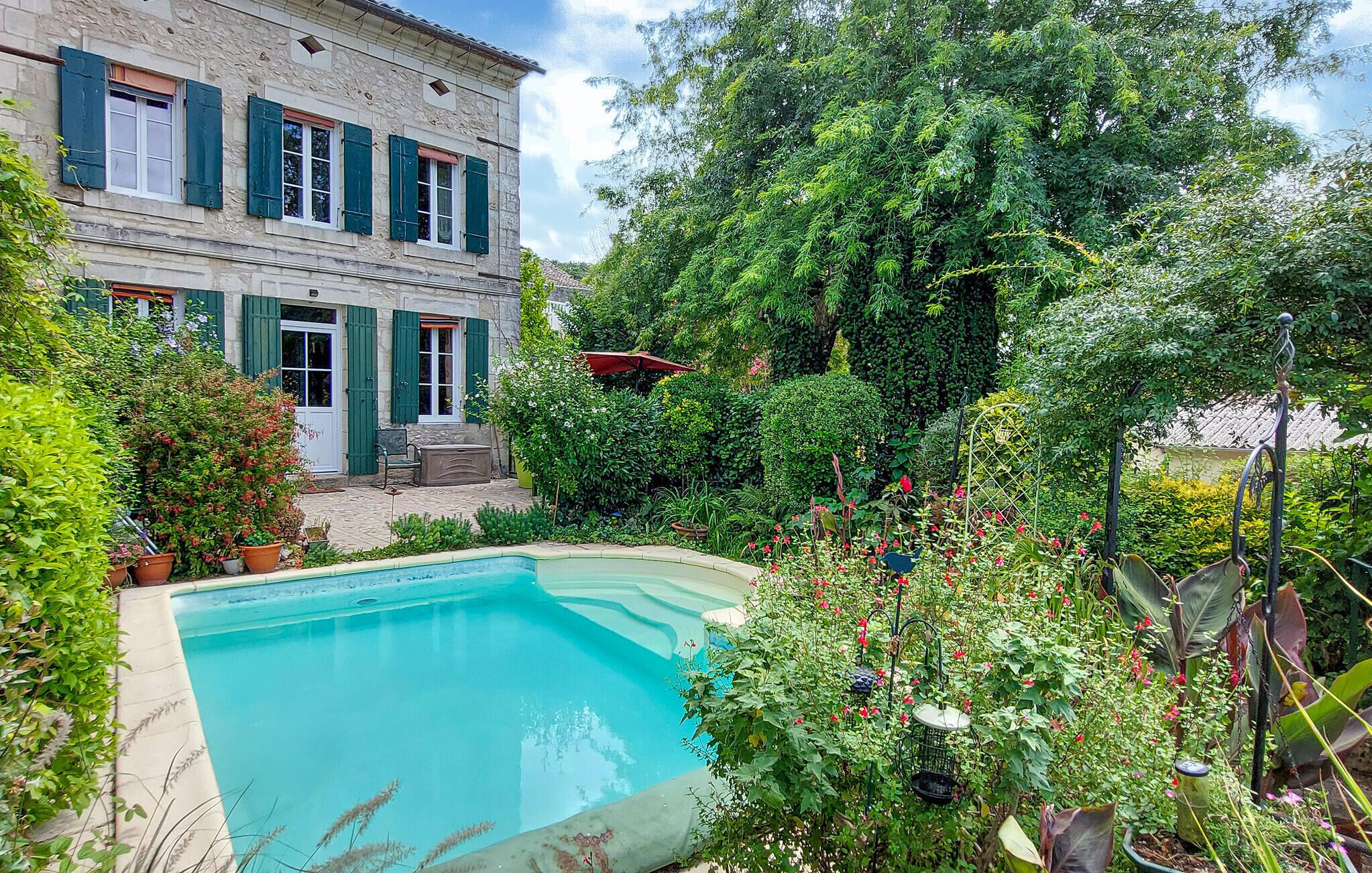 Una piscina circondata da vegetazione lussureggiante e un edificio in pietra con persiane verdi.