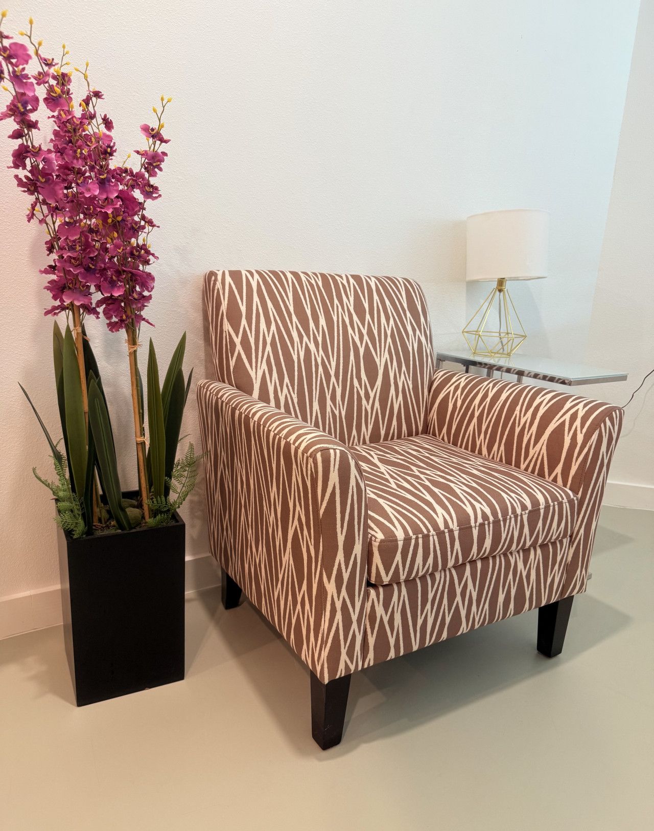 A modern armchair with a geometric pattern, positioned next to a black planter with green foliage and purple orchids.