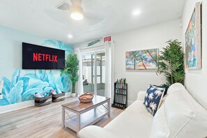 Un salon moderne avec un téléviseur à écran plat affichant le logo « NETFLIX », un canapé blanc, une table basse en bois et une porte marquée « SORTIE ».