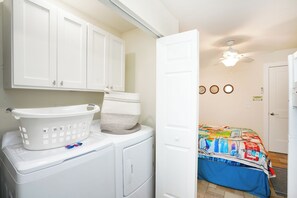 A laundry area with a washing machine, a dryer, and a basket. There is a bed with a colorful bedspread in the adjacent room.