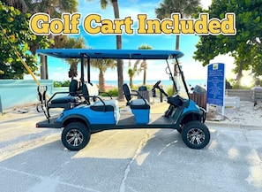 En golfbil med blåt tag er parkeret på en belagt plads med palmer og en strand i baggrunden.
