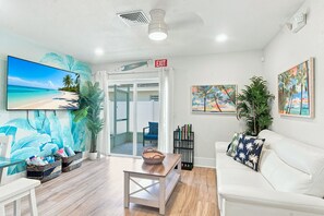 A room with a beach-themed mural, a white sofa, a small table, and a TV screen displaying a beach scene. - 4bd Duplex 1 blk from bch, pool Hot Tub, golf cart (Indian Rocks Beach)