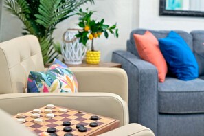A living room with a beige sofa, a colorful cushion, a grey couch with blue and pink pillows, a small table with a plant and a vase, and a wooden board game. - 4bd Duplex 1 blk from bch, pool Hot Tub, golf cart (Indian Rocks Beach)