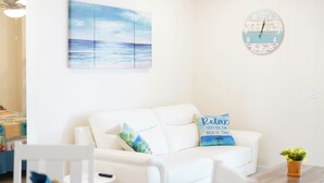 Un salon avec un canapé blanc, une horloge, un tableau représentant une plage et une table sur laquelle se trouve une plante.