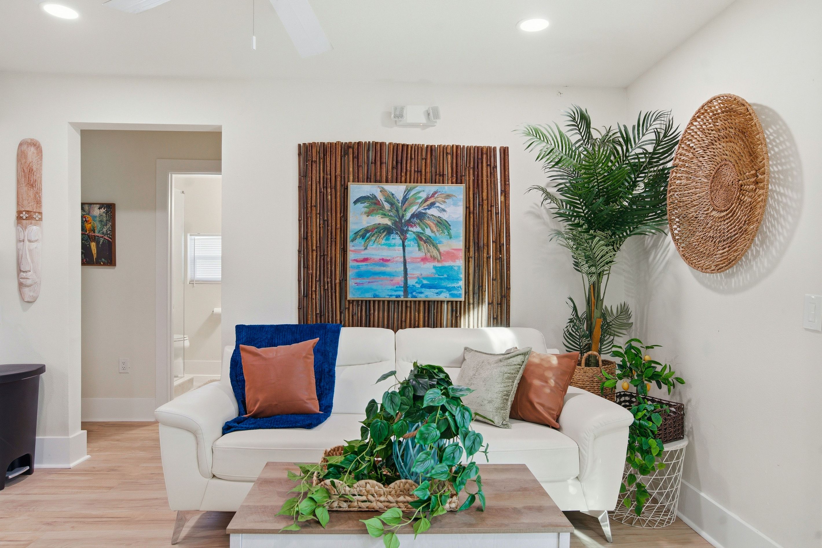Un salón con un sofá blanco, una mesa de centro con plantas, un cuadro de una palmera y una pieza de arte en la pared hecha con fibras naturales.