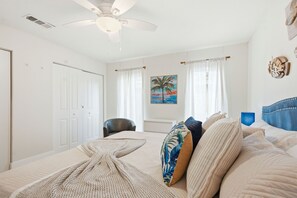 A bedroom with a bed, a ceiling fan, a chair, and a painting of a palm tree.