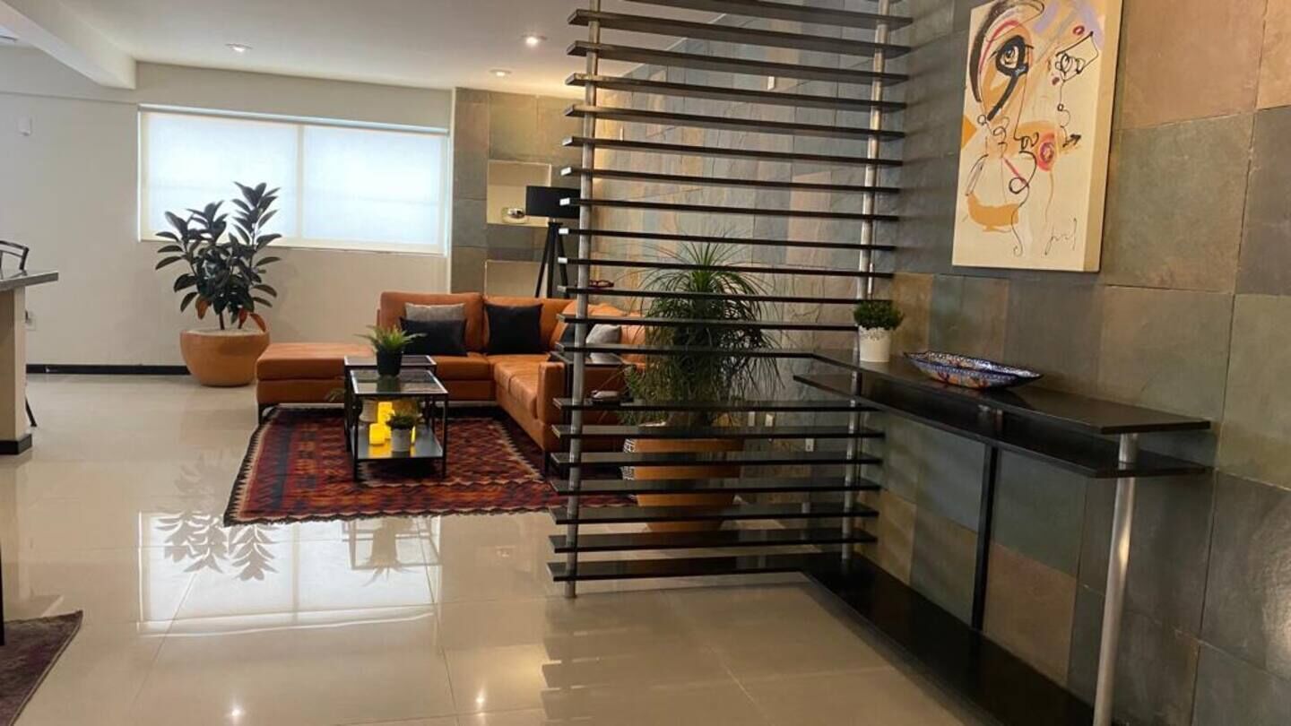 A modern living room with a sofa, coffee table, and plants. There is a staircase with a railing and a painting on the wall.