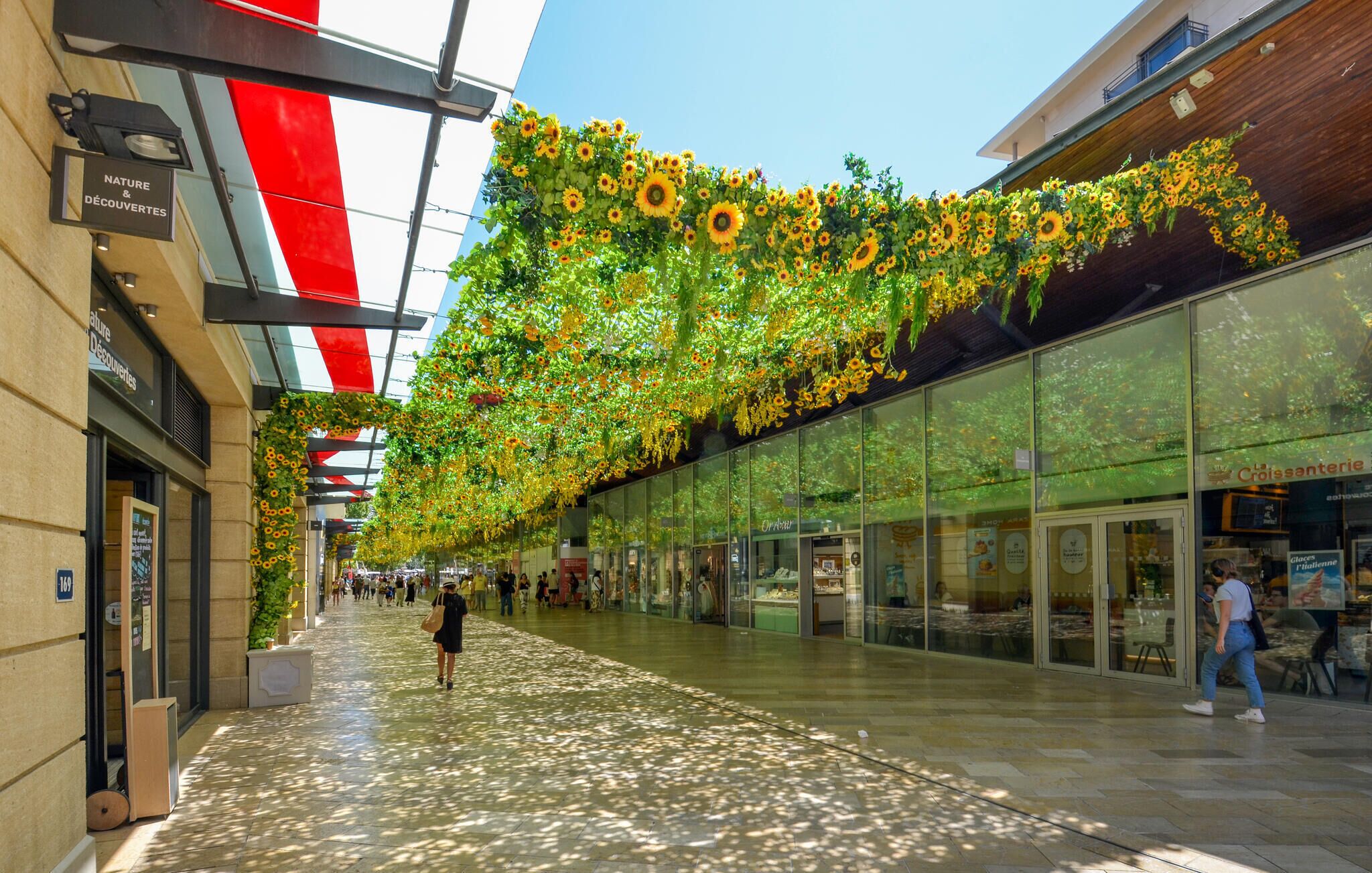 A covered walkway with a ceiling adorned with sunflowers, glass storefronts, and a red and white striped structure.
