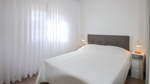 A bedroom with a bed, curtains, a nightstand with a lamp, and a drawer unit.