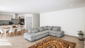 A modern living room with a grey sectional sofa, a dining area with white chairs, and a kitchen with white cabinets and a patterned backsplash.