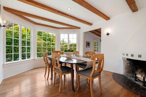 A dining room with a round table and wooden chairs, large windows with a view of greenery, and a fireplace. - Monte Sagrado (Montecito)