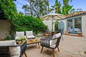 An outdoor patio with wicker furniture, a table with a bottle and fruit, and a large umbrella.