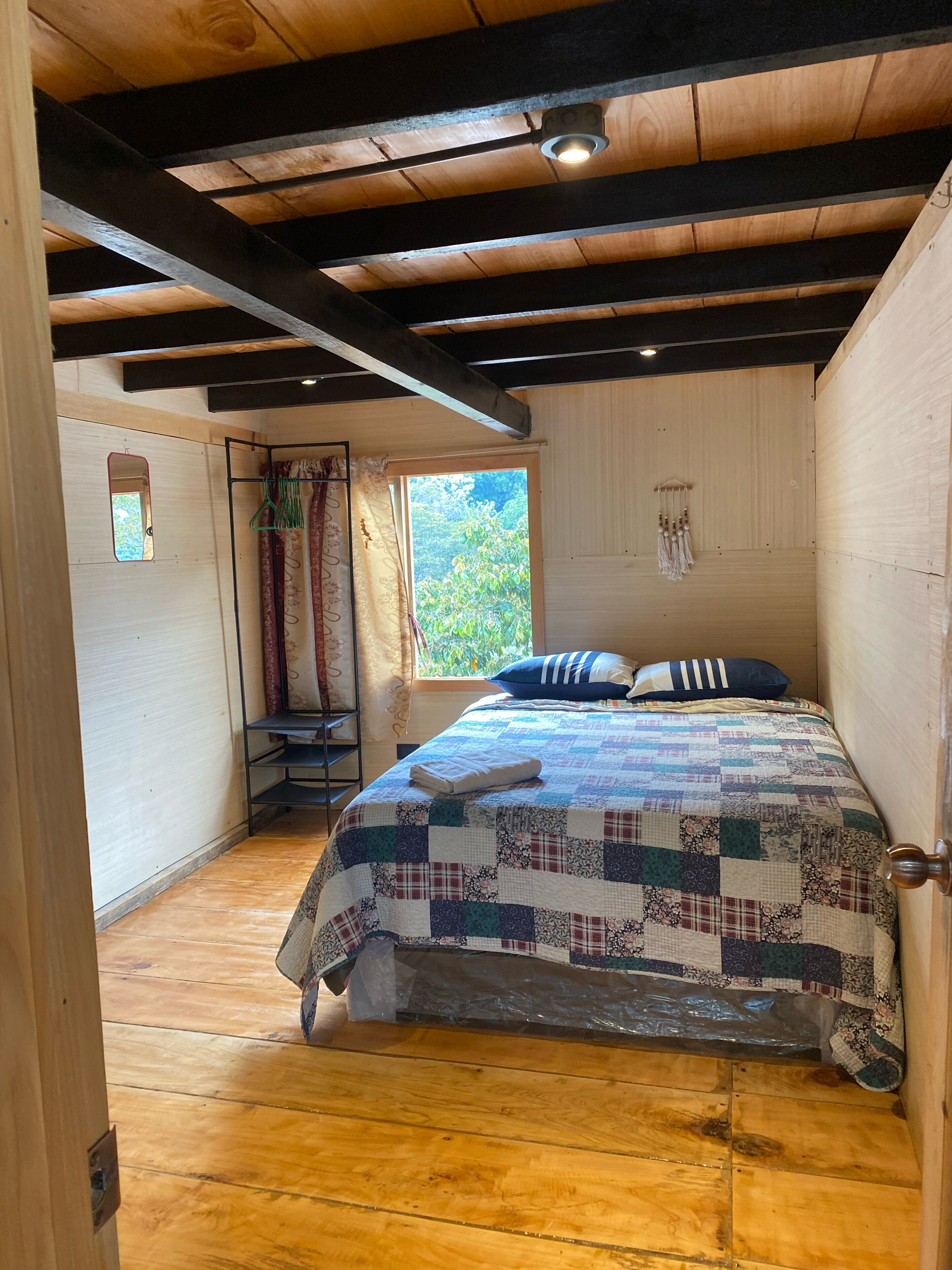 A bedroom with a wooden floor, a bed with a quilt, a window with curtains, and a mirror on the wall.