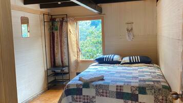 A bedroom with a wooden floor, a bed with a quilt, a window with curtains, and a mirror on the wall.