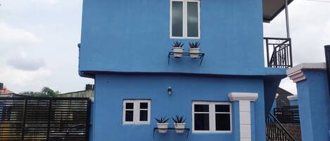 Un edificio de dos pisos con fachada azul, una puerta blanca y dos ventanas con plantas en macetas.