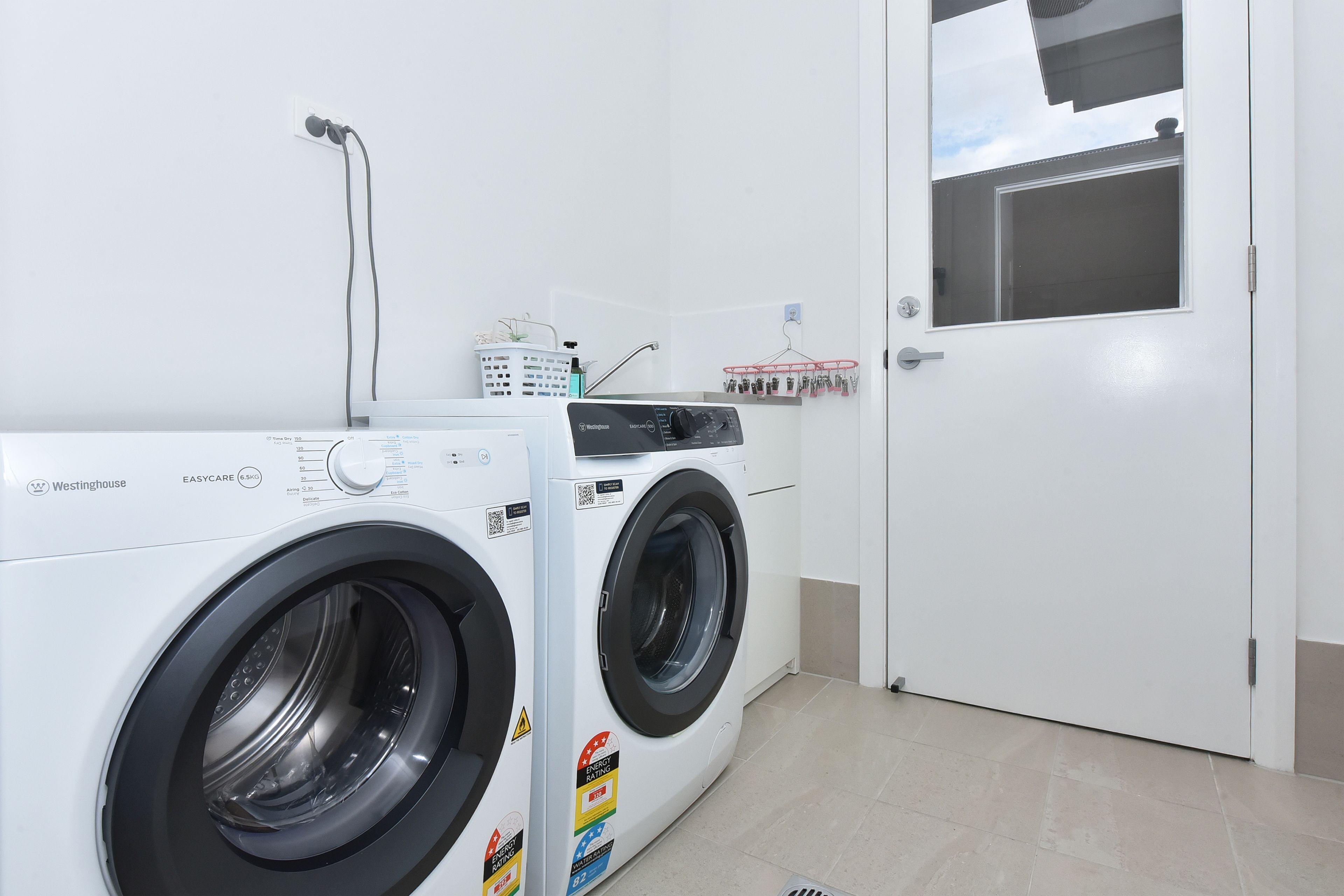 A laundry area with two white washing machines, a white door, and a window with a view of a building.