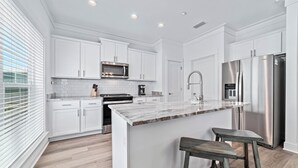 A modern kitchen with white cabinetry, a stainless steel refrigerator, and a marble countertop island. - Laughing Gull at Summer Salt | Zero Entry Pool | Beach Access | Lake Fishing | My Beach Getaways (Orange Beach)