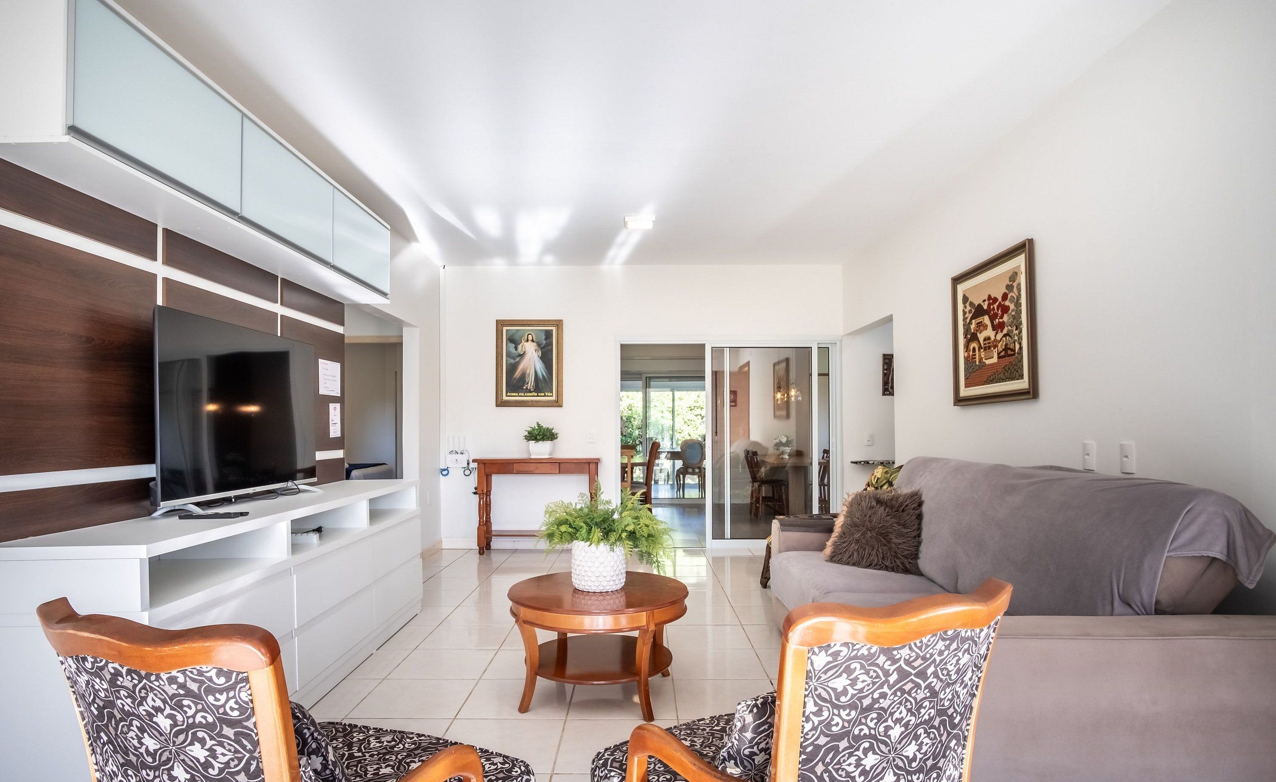 Uma sala de estar moderna com televisão de tela plana, um sofá, poltronas estampadas, uma mesa de madeira redonda com uma planta e obras de arte nas paredes.