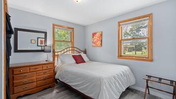 A bedroom with a wooden bed, a dresser, a mirror, and two windows with a view of trees and a building.