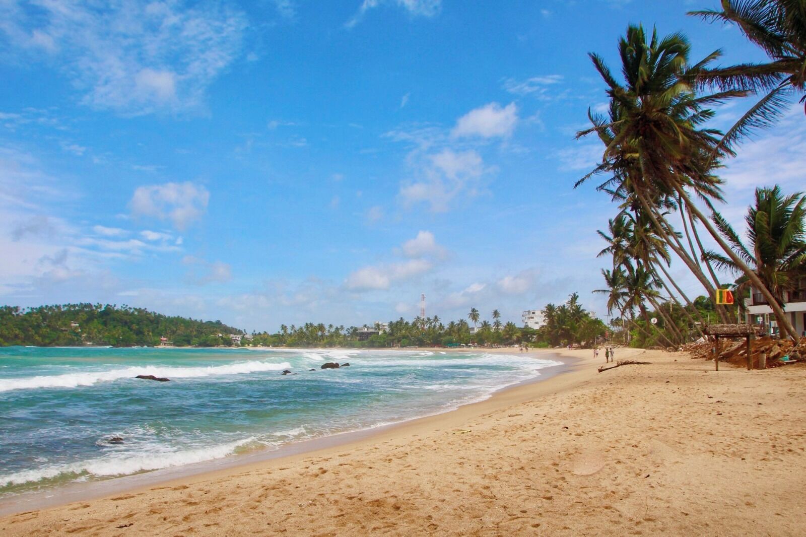 Uma praia com águas turquesa cristalinas, palmeiras e uma linha de costa distante com edifícios.