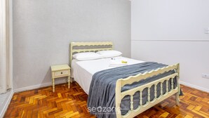 Une chambre avec un parquet en bois disposé en motif «chevrons», un lit avec un matelas blanc et une couverture grise, une petite table de chevet avec une lampe, et un mur blanc uni.