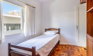 Una habitación pequeña con cama individual, cabecera de madera, ventana con cortinas blancas y piso de madera con patrón de espiga.