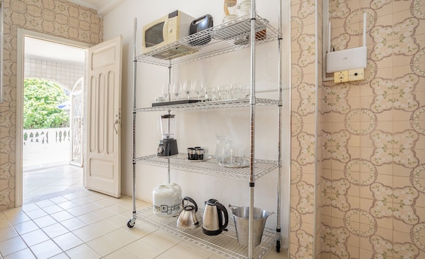 Una cocina con una estantería metálica que contiene utensilios de cocina y electrodomésticos, suelo de baldosas y una pared con diseño.