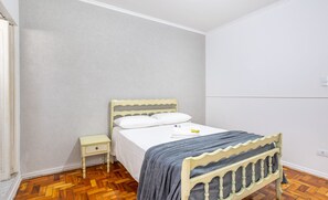 Une chambre avec un parquet en bois disposé en motif «chevrons», un lit avec un matelas blanc et une couverture grise, une petite table de chevet avec une lampe, et un mur blanc uni.