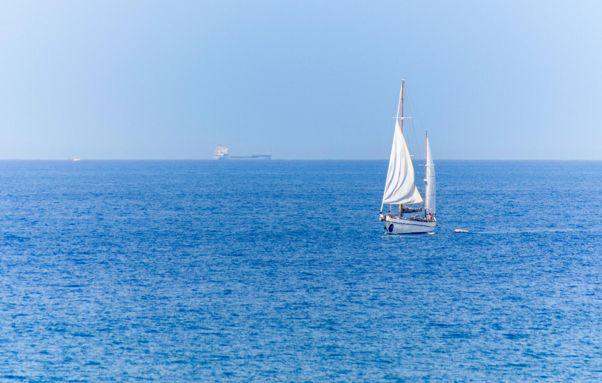 A sailboat on a calm sea with a distant ship in the background.