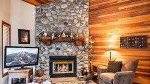 Una sala de estar acogedora con chimenea de piedra, paredes de madera, televisor y una zona de estar confortable.