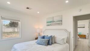 A bedroom with a large bed, a window, a bedside table with a lamp, and a door leading to another room. - 7 Mi Huntington Beach: Family Home w/ Patio! (Midway City)