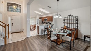 A spacious dining area with a wooden dining table, chairs, and a glass-top table. A kitchen with wooden cabinets and a microwave is visible in the background.