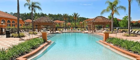 A swimming pool with lounge chairs and gazebos, surrounded by palm trees and buildings with red-tiled roofs.