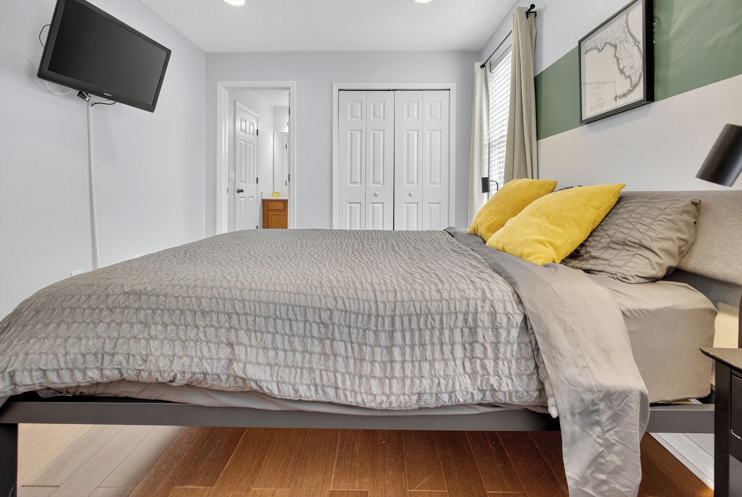 A bedroom with a bed, a flat-screen TV mounted on the wall, a framed map, and a door in the background.