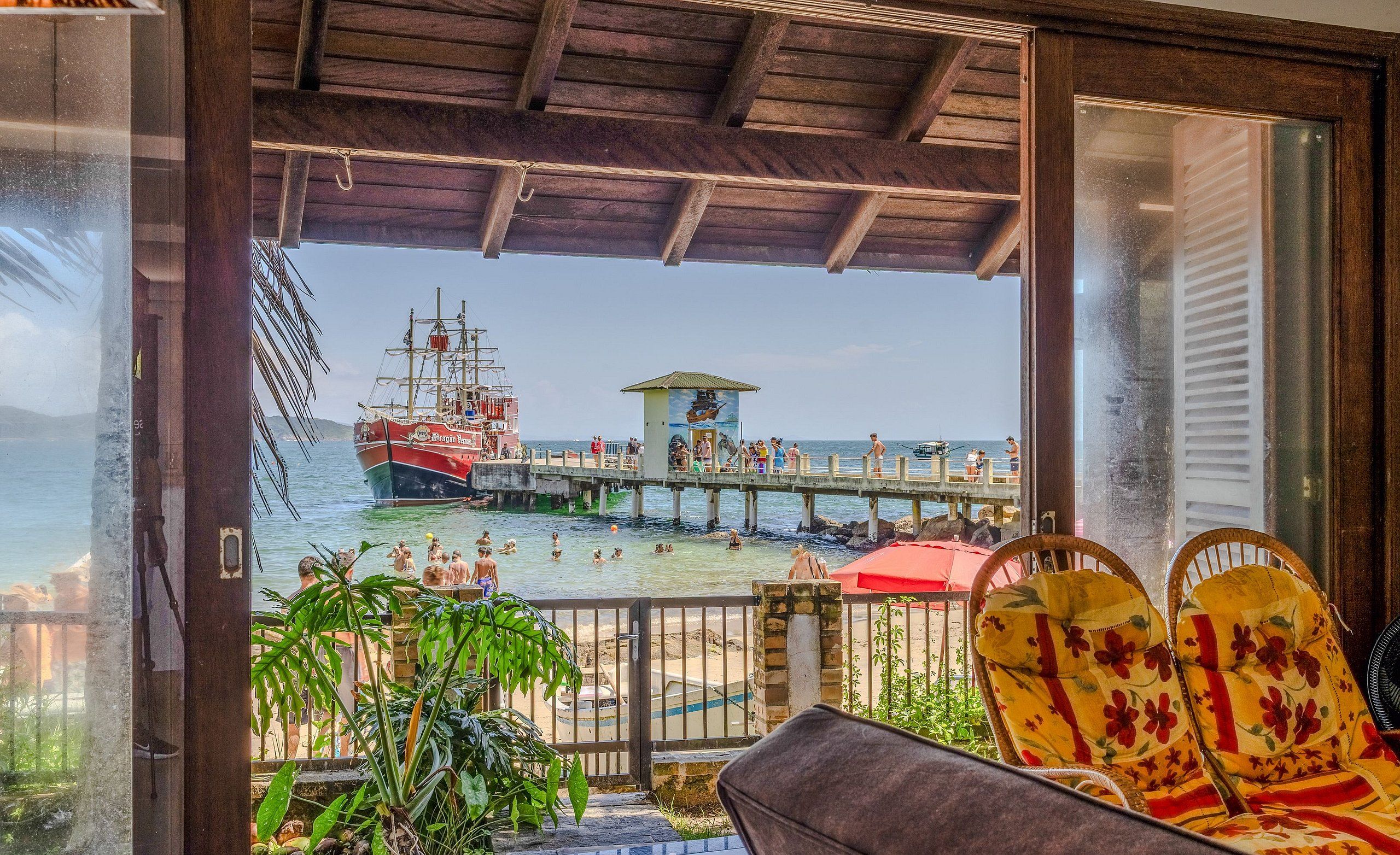 A beach view from a covered area with a wooden ceiling, a lifeguard tower, and a pier with a ship.