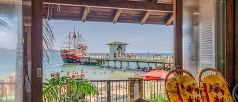 A beach view from a covered area with a wooden ceiling, a lifeguard tower, and a pier with a ship.