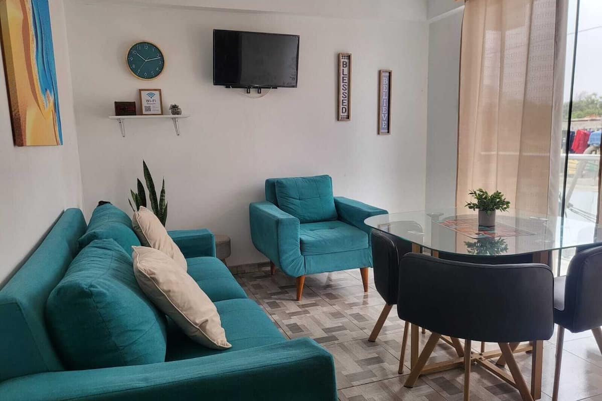 A living room with teal sofas, a glass table, and a TV mounted on the wall.